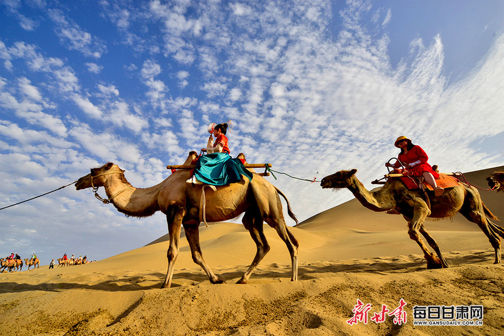 游客骑骆驼畅游鸣沙山,体验敦煌壮丽的大漠风景和浓郁的丝路风情.
